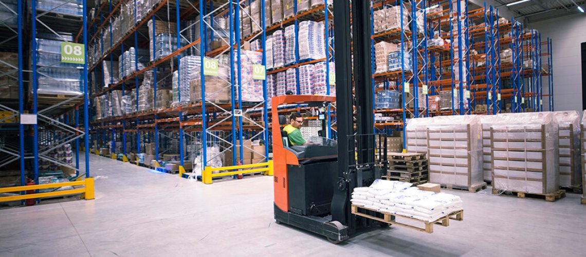 Worker operating forklift machine and relocating goods in large warehouse center.